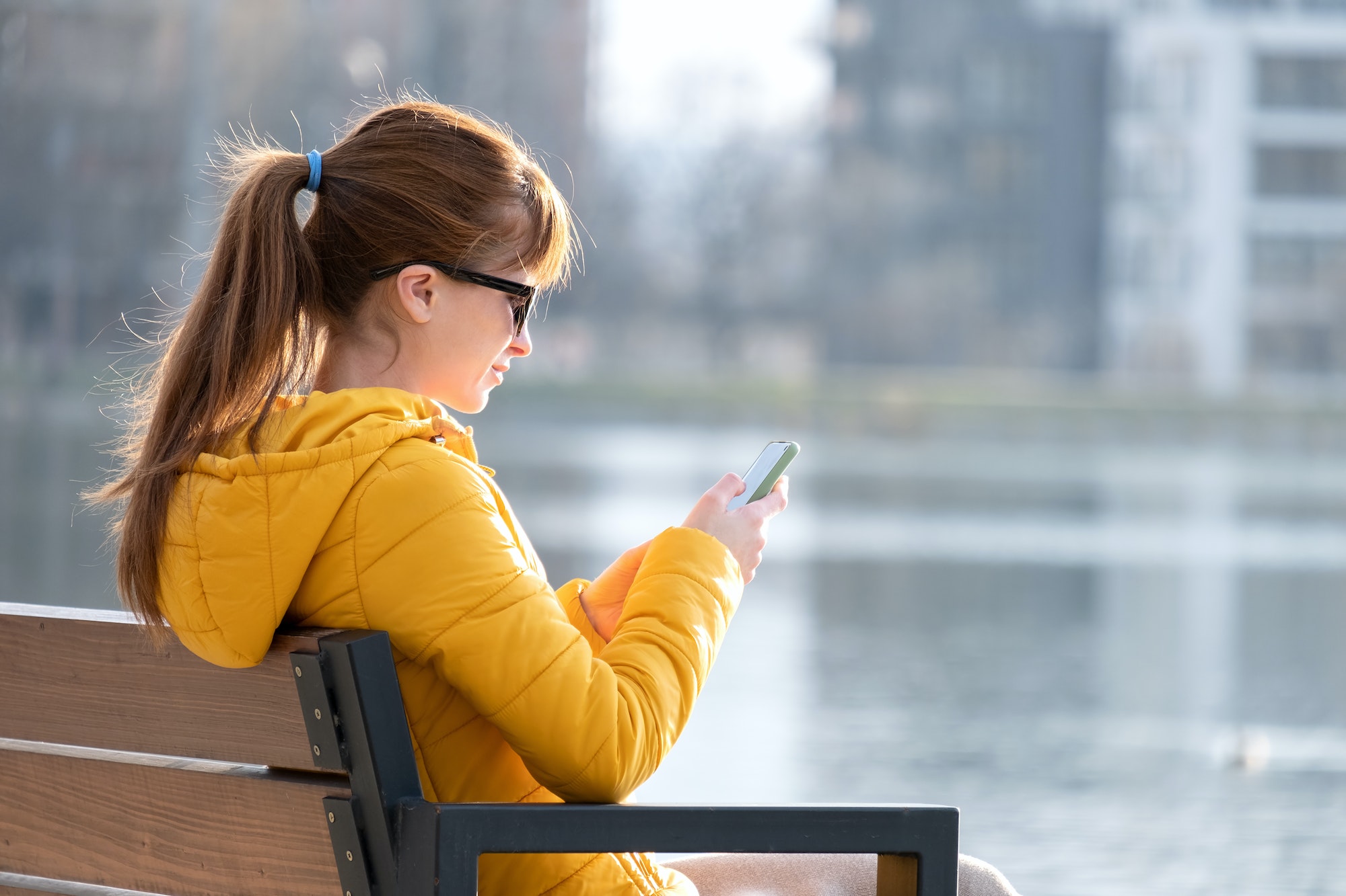 Young pretty woman sitting on a park bench browsing her smartphone outdoors on warm autumn day
