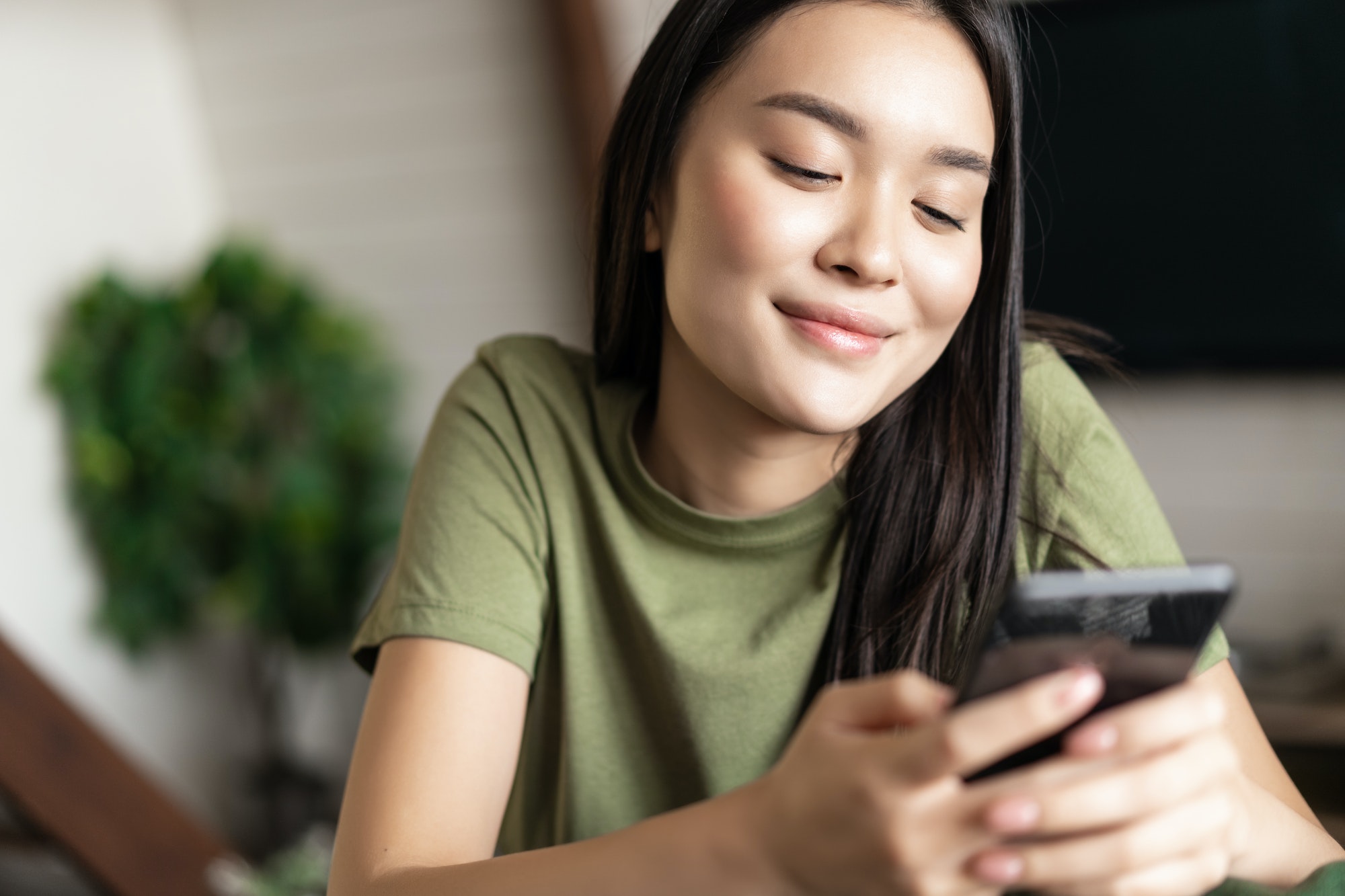 Smiling korean woman looking at mobile phone chat app with dreamy face, sitting at home on couch